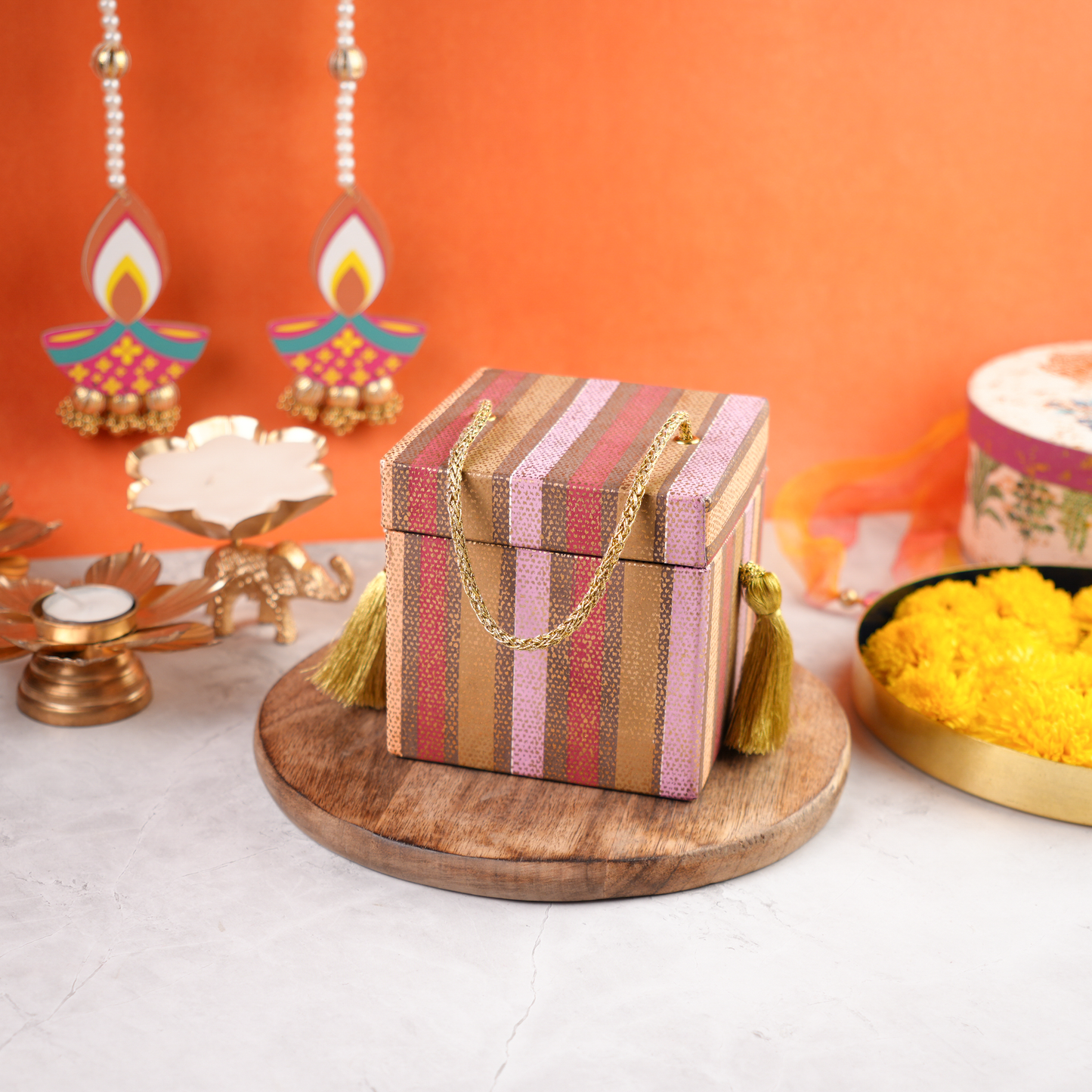 An angled view of a closed reusable striped trunk box decorated with golden tassels, placed on a wooden platform with beautiful décor in the background.