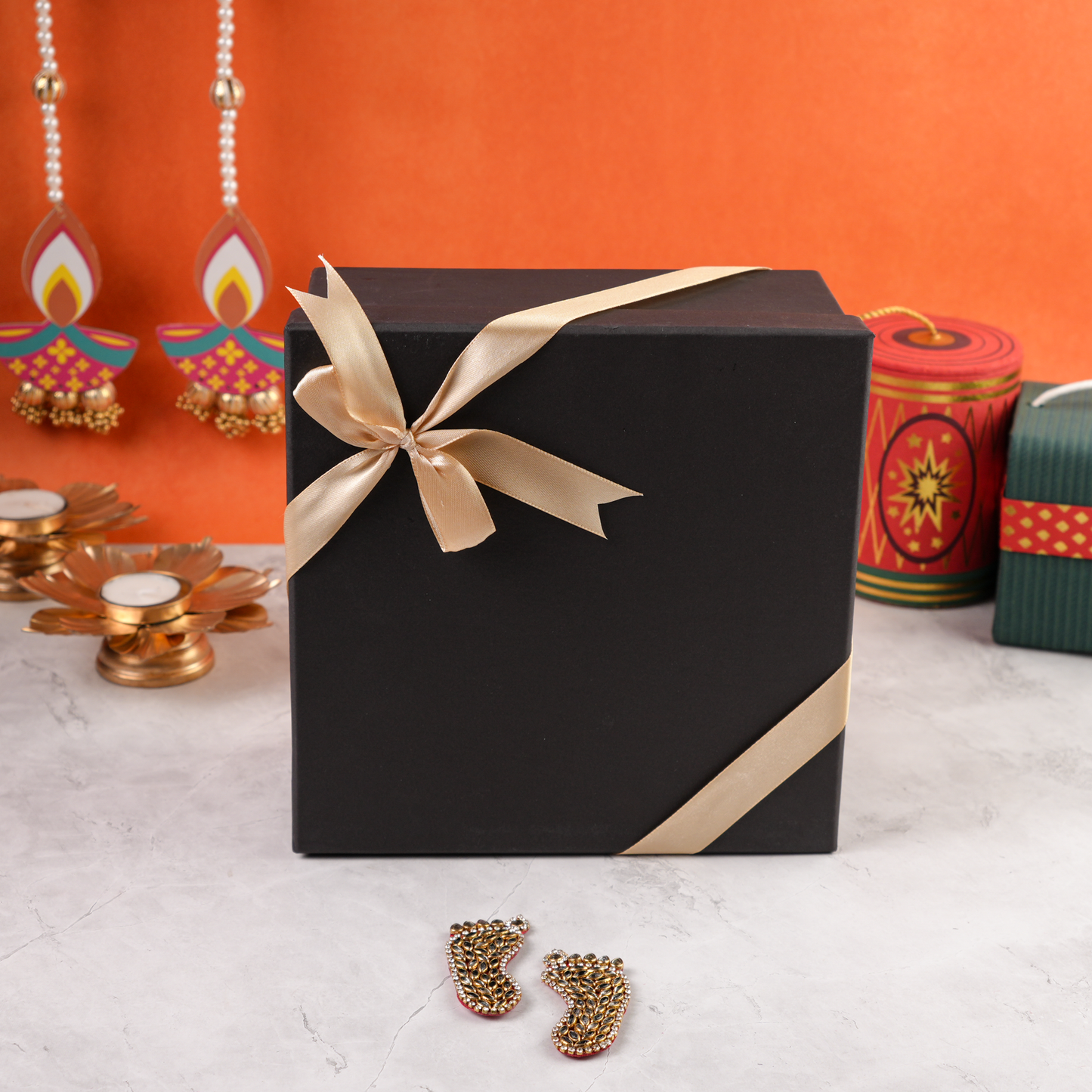 Closed black Diwali gift box with gold ribbon, decorative diyas behind and Laxmi Charan in front.