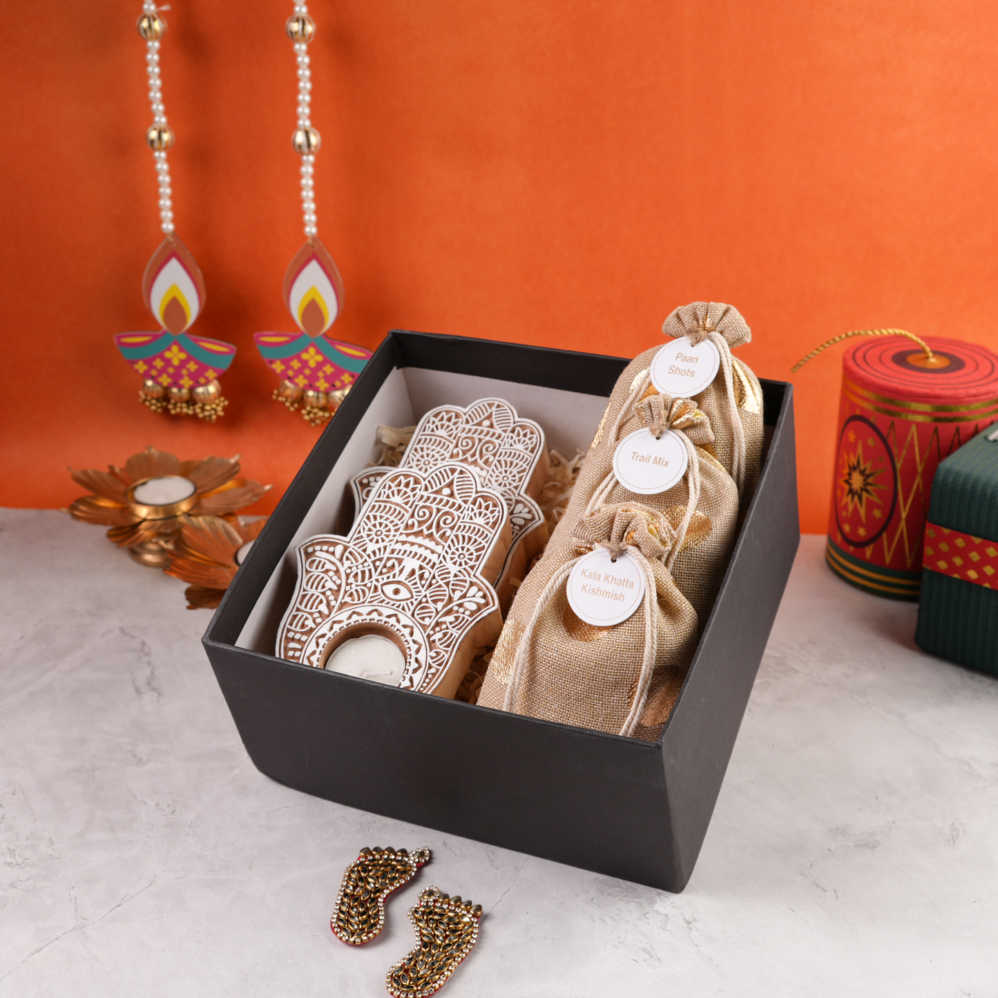 Open black gift box at an angle with hand-pattern tealight holders and festive jute pouches, Diwali backdrop.
