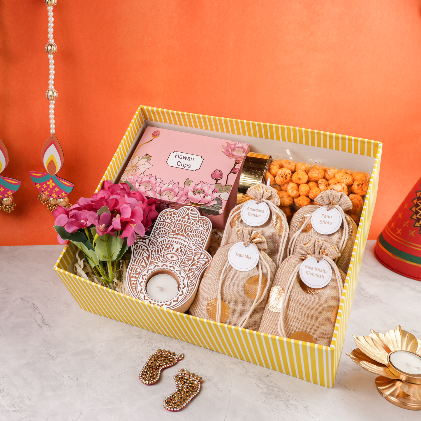 Angled view of open yellow striped Bagh & Blossom Diwali hamper displaying potlis, golden tealight holder, and pink flower bunch with festive accents.