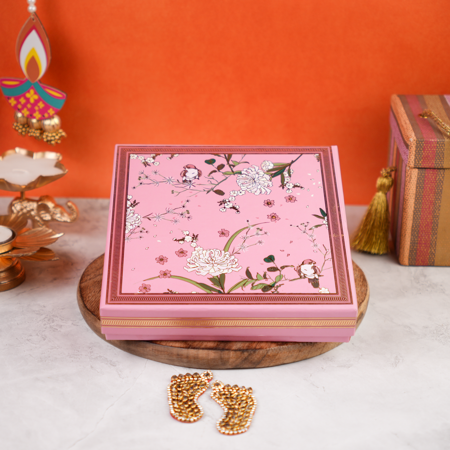 Closed front close-up of the Floral Budget Diwali Gift Box—pink floral lid with bird-and-bloom motif—placed on a wooden tray against an orange festive backdrop.