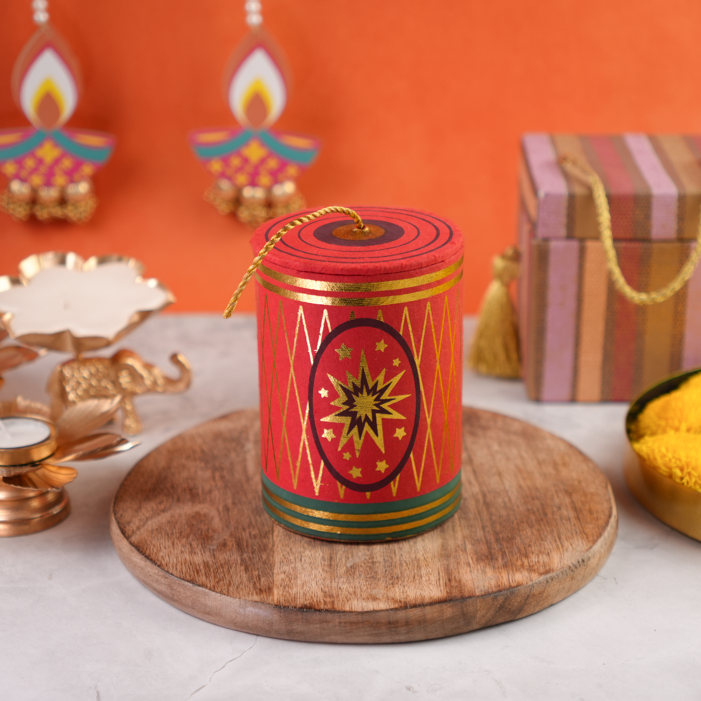 Close-up of Rangeela Chilly Bomb Diwali gift box with golden rope handle on a wooden base and marigold flowers in background