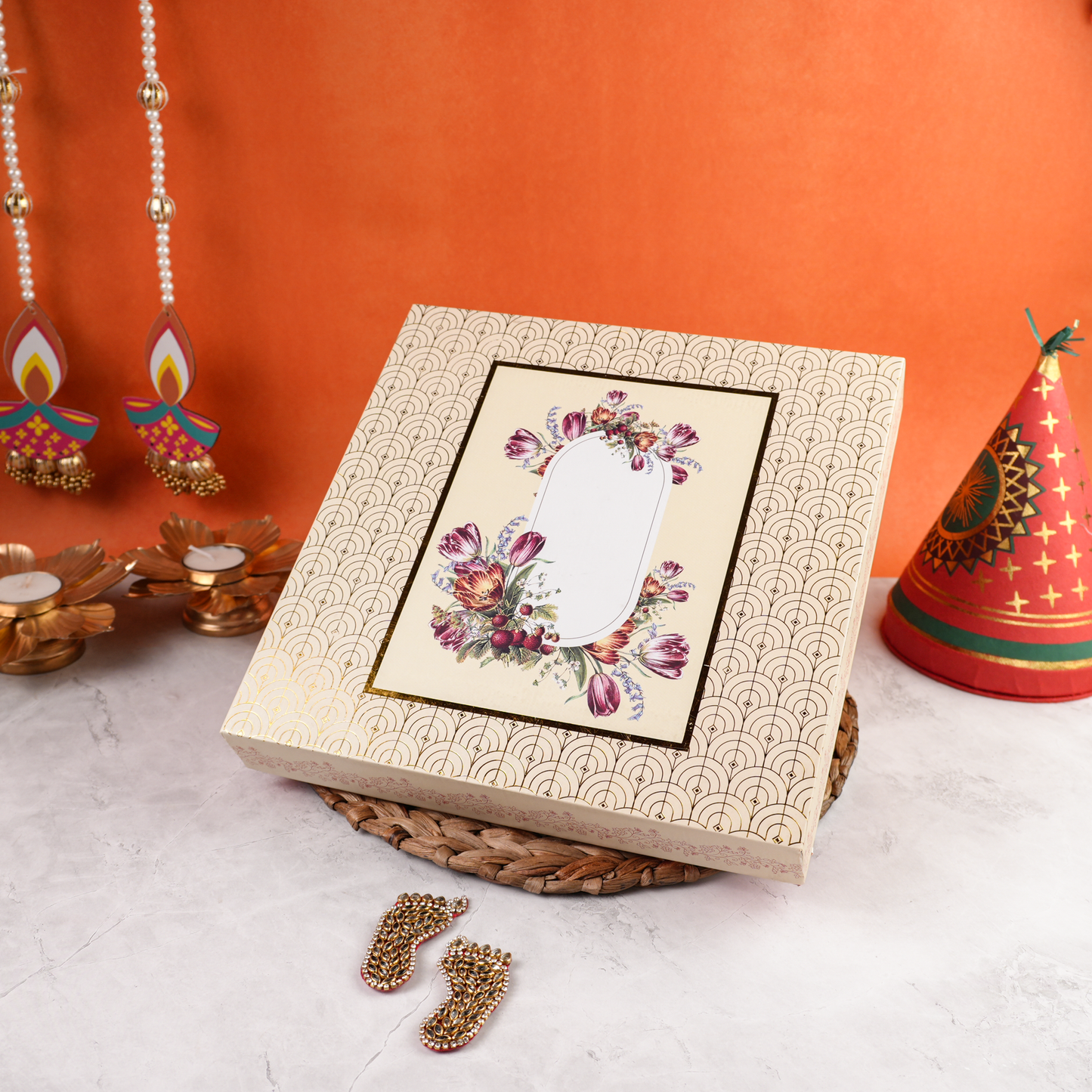 Cream rigid Diwali gift box with gold-foil pattern and floral panel, closed, with festive backdrop and Laxmi Charan.