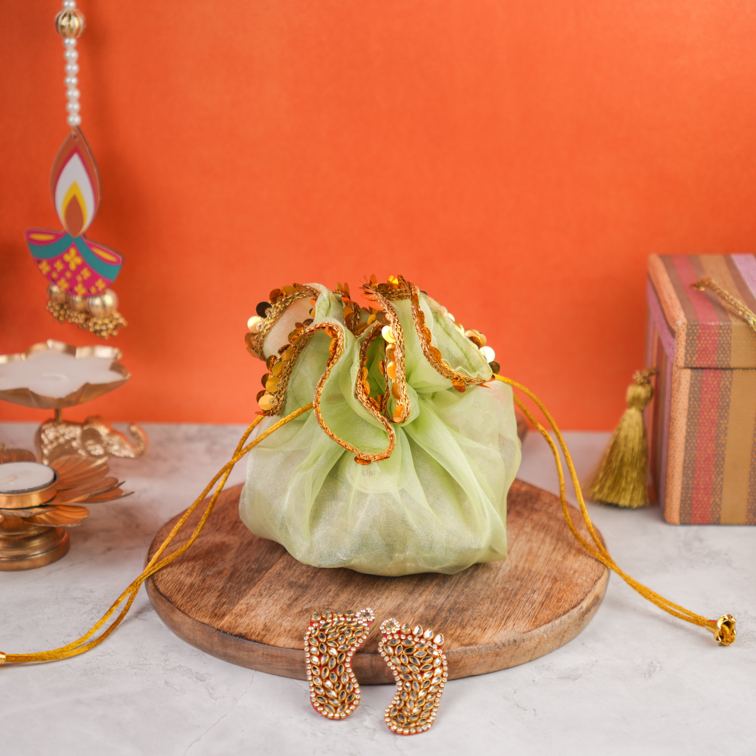 A light green organza drawstring potli decorated with golden lace and pearl embellishments, placed on a wooden board, set against an orange festive background.