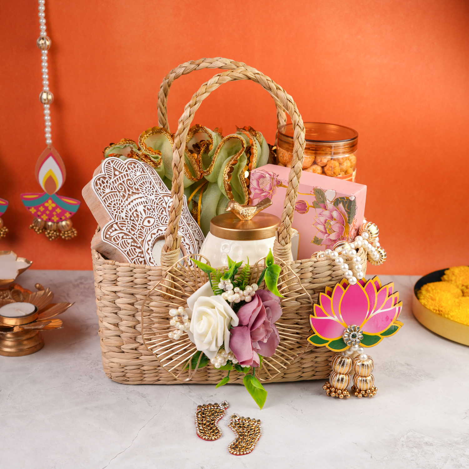 Woven kauna grass gift basket filled with carved wooden candles, a decorative floral bunch, a pink gift box, and a snack jar, displayed with diyas and festive decor against an orange background.