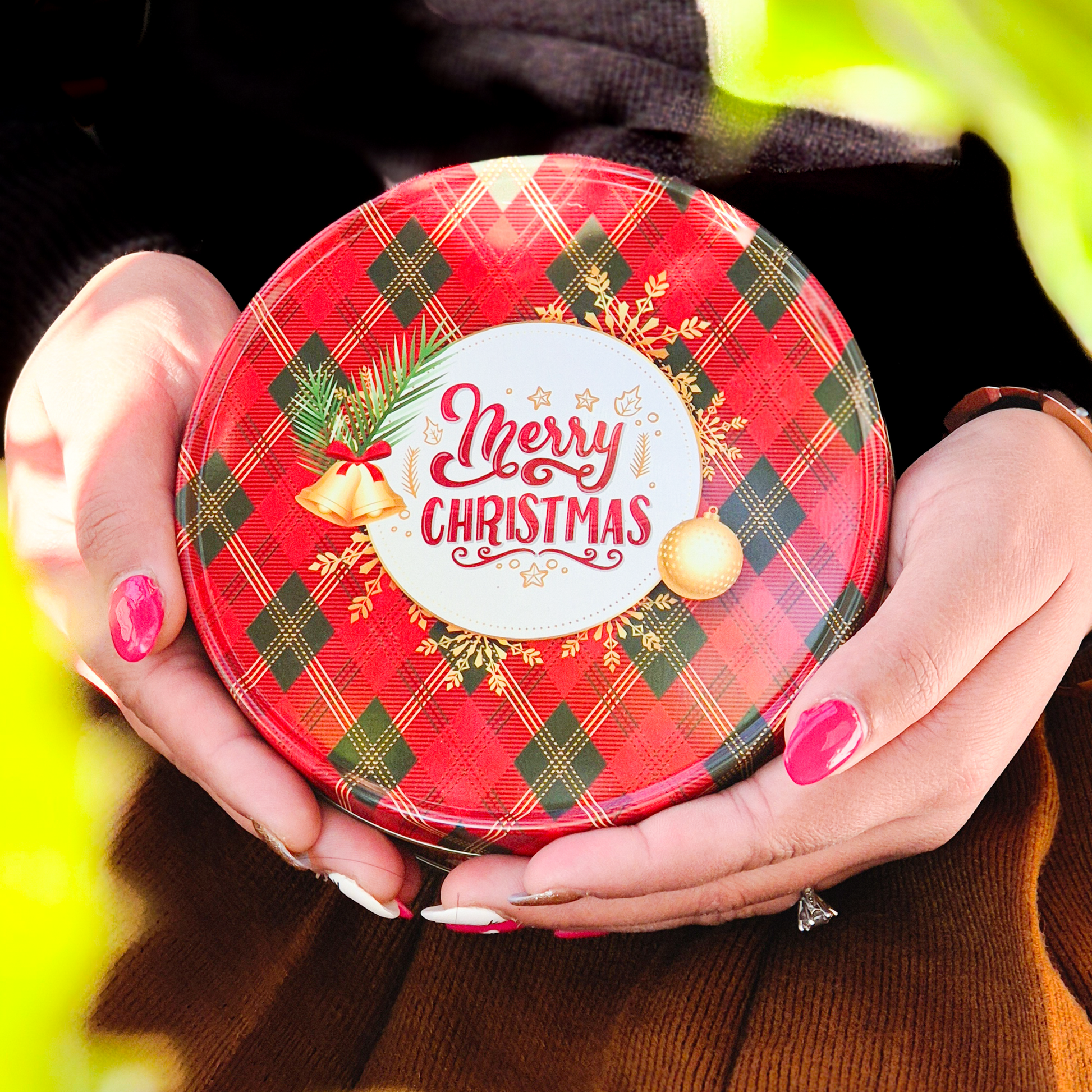 Hands holding a round red and green tartan-patterned Christmas tin decorated with bells, pine leaves, ornaments, and the text “Merry Christmas.”