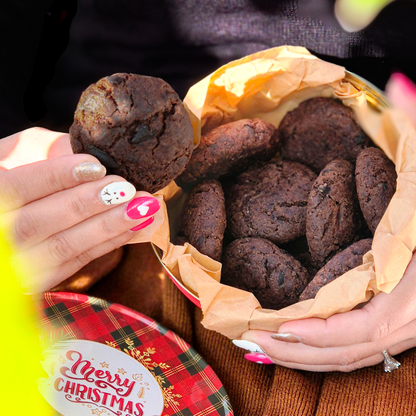 Hand holding a chocolate chip cookie over a Christmas-themed tin filled with soft-baked cookies layered in parchment paper.