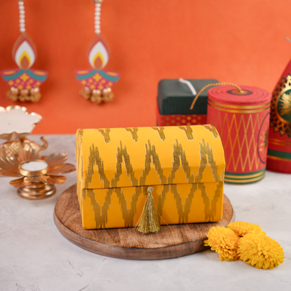 Ikat Diwali trunk hamper, saffron-and-gold dome lid with tassel, festive props in the background.