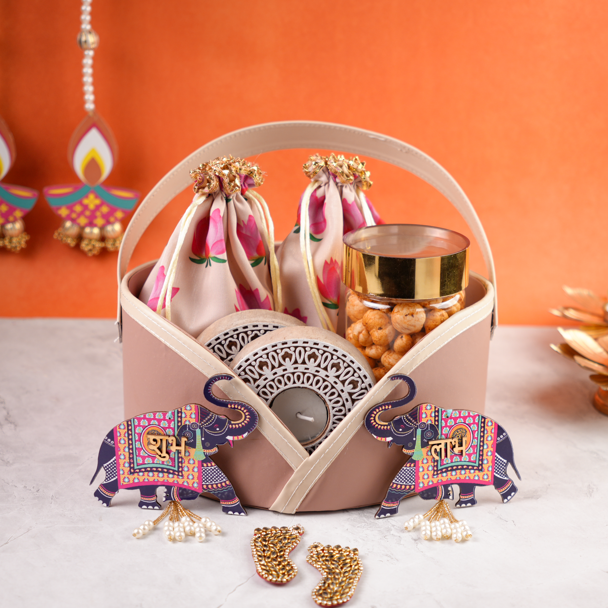 Zoomed in view of pastel pink wedding gift basket with lotus print pouches, tealight holder, and snacks against orange backdrop.