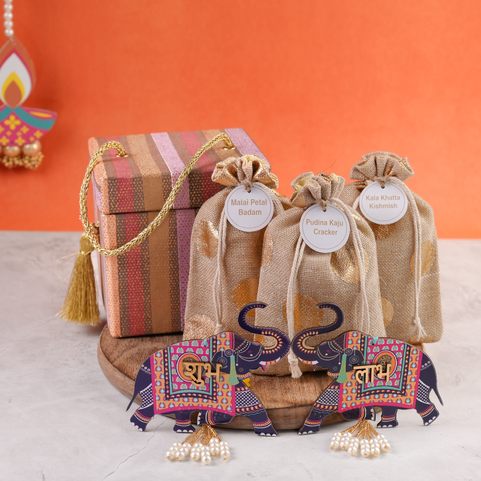 A premium striped gift trunk displayed open with three jute potlis filled with assorted treats, featuring golden tassel details and arranged on a wooden platform.