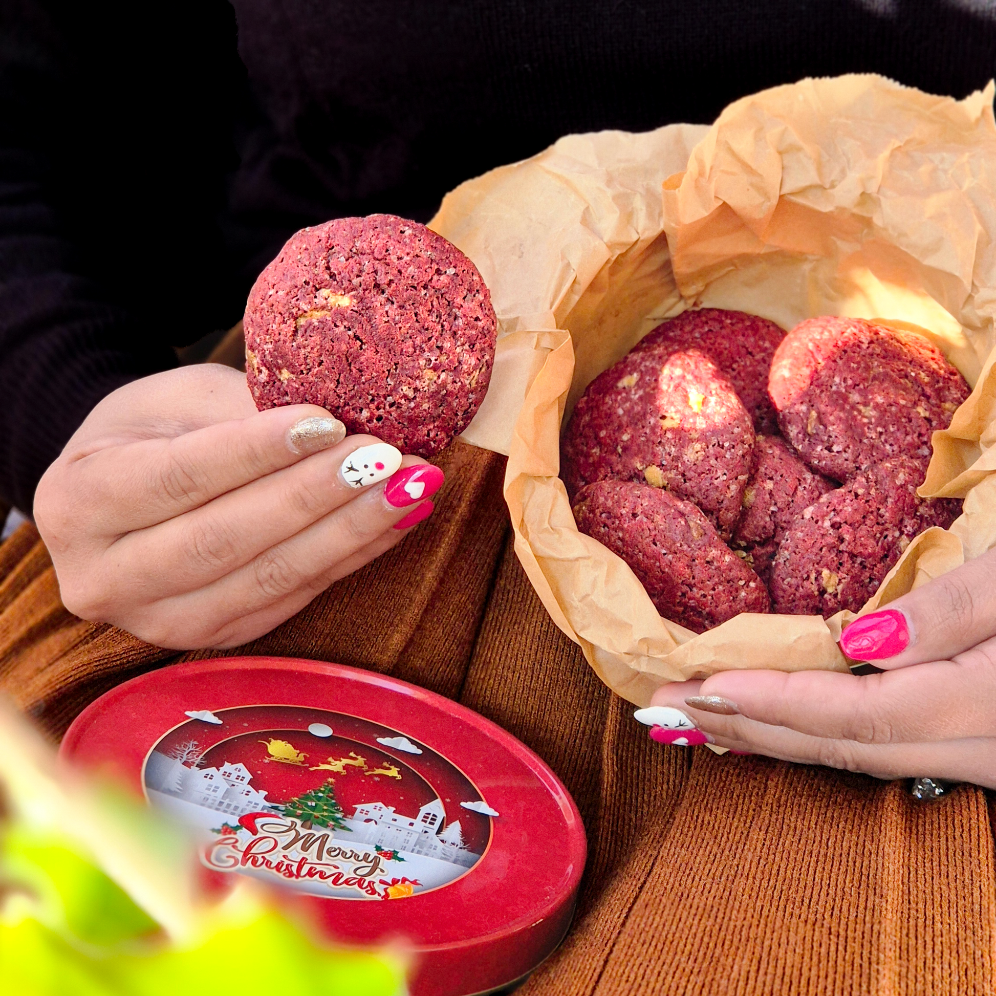 Hand holding a red velvet cookie over a Christmas-themed tin filled with soft-baked cookies layered in parchment paper.